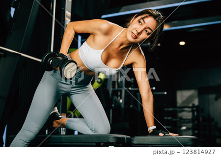 Asian woman lifting dumbbells on a bench in a fitness gym, happy young female performing strength training exercises with dumbbells in a gym, weight training for a healthy lifestyle concept Asian woman lifting dumbbells on a bench in a fitness gym, happy young female performing strength training exercises with dumbbells in a gym, weight training for a healthy lifestyle concept 123814927