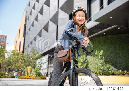 A cheerful Asian businesswoman suited up and helmeted stands with her bicycle ready for the morning office commute. This image captures the essence of a modern business commuter. A cheerful Asian businesswoman suited up and helmeted stands with her bicycle ready for the morning office commute. This image captures the essence of a modern business commuter. 123814934