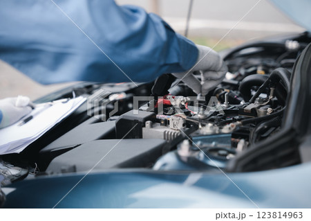 Focused Asian mechanic with a clipboard inspecting car engine in garage. Expertise in car service and repair working on the challenge. Technician ensuring safety and control technology-driven. 123814963