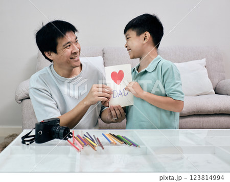 Smiling young boy presents handmade Father's Day card with red heart and "DAD" written on it to his father, sharing heartfelt moment at home 123814994