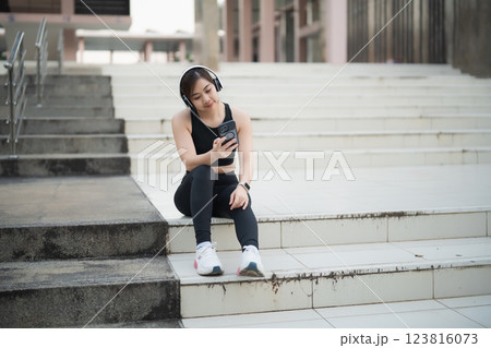 Asian Young woman wearing headphones sits on steps while checking her phone, enjoying music or podcasts in an outdoor urban setting Asian Young woman wearing headphones sits on steps while checking her phone, enjoying music or podcasts in an outdoor urban setting 123816073
