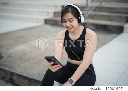 Asian Young woman enjoying music on headphones while checking phone outdoors in urban setting with active lifestyle and casual sportswear Asian Young woman enjoying music on headphones while checking phone outdoors in urban setting with active lifestyle and casual sportswear 123816074