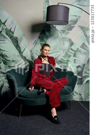 Stylish woman poses in burgundy outfit on green couch with tropical backdrop in modern setting Stylish woman poses in burgundy outfit on green couch with tropical backdrop in modern setting 123817735