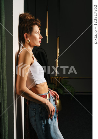 A thin woman in jeans and top stands in profile, leaning her back against the wall 123817852
