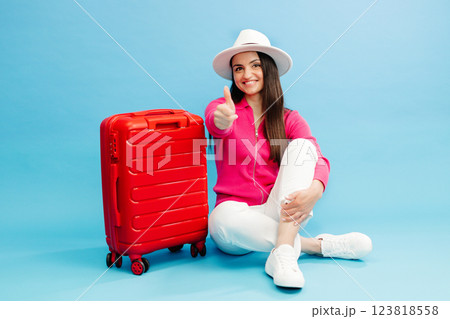 Happy young tourist woman sitting at the suitcase, showing finger up gesture and going to travel on holidays isolated on blue background. Happy young tourist woman sitting at the suitcase, showing finger up gesture and going to travel on holidays isolated on blue background. 123818558