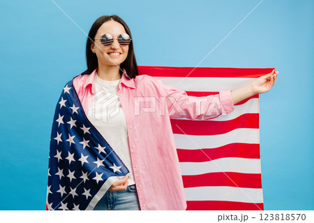 Young smiling happy woman wears casual clothes holding american flag and looking to the camera isolated on blue color background. Young smiling happy woman wears casual clothes holding american flag and looking to the camera isolated on blue color background. 123818570