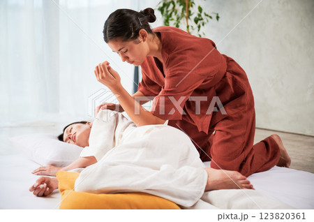 Masseuse making thai yoga massage. Woman therapist doing traditional massage treatment for female patient using elbow. Client lying on mat, meditating, enjoying hand massage in calm atmosphere. 123820361