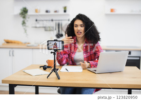 African woman in casual outfit recording video content in modern kitchen using smartphone tripod. Engaging with viewers through online platform. Creating home-based work or hobby project. 123820950