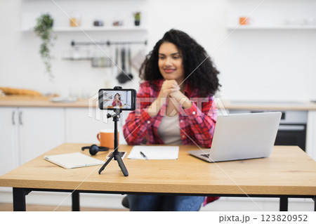 African woman in her thirties recording video blog at home using smartphone. Woman in casual outfit seated at table with laptop, notepad, and coffee. Expression indicates engagement and communication. 123820952