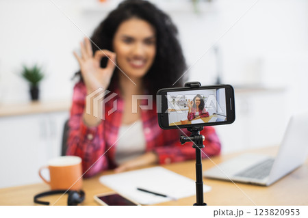 African woman in her 20s recording vlog using smartphone, wearing casual clothes, sitting in home office with coffee, papers, and laptop on table, creating engaging content for social media platforms. 123820953
