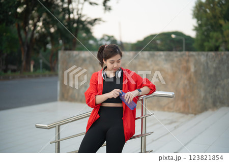 Asian Young woman in red jacket adjusts water bottle by railing, wearing headphones, active lifestyle, urban environment, enjoying outdoor exercise, healthy habits Asian Young woman in red jacket adjusts water bottle by railing, wearing headphones, active lifestyle, urban environment, enjoying outdoor exercise, healthy habits 123821854