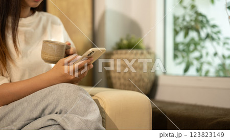 Close up of a woman holding a warm coffee mug and using smartphone in a living room 123823149