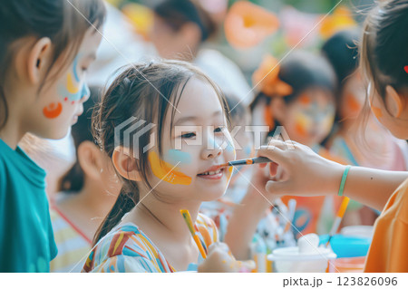 Young girl smiling while getting her face painted by a friend at an outdoor party 123826096