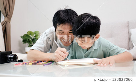 Father and son smiling while drawing together at home, using colored pencils and sketchbook on glass table in cozy living room Father and son smiling while drawing together at home, using colored pencils and sketchbook on glass table in cozy living room 123827060