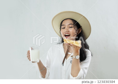 Young Asian woman in straw hat passionately eating bread in her hand and in her hand holding a glass of milk, woman breakfast concept, isolated white background 123830384