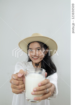 portrait Young Asian woman wearing hat with white top pushing a glass of milk to the camera to tell drink milk, isolated white background 123830509