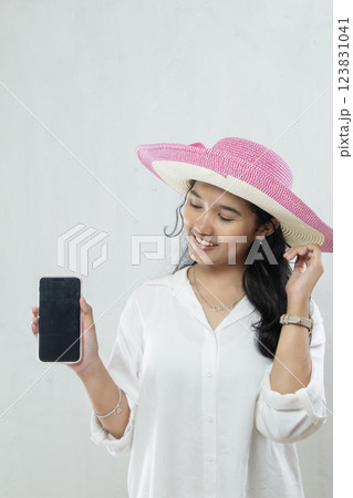 portrait of young Asian woman wearing straw hat, all white outfit showing black screen of mobile phone, used for travel advertising via app, mobile phone mockup 123831041