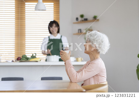 キッチンで料理をする30代の女性とテーブルでお茶を飲むシニア女性 家事代行イメージ 123831221