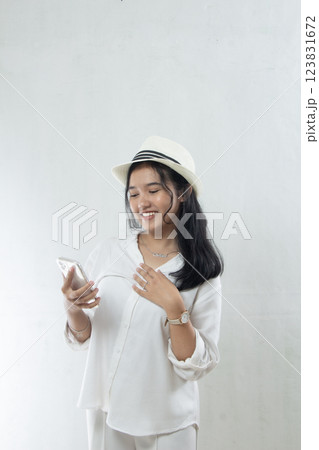 Beautiful young Asian woman tourist in straw hat, all-white suit standing smiling shyly sending message from mobile phone in her hand, isolated on white Beautiful young Asian woman tourist in straw hat, all-white suit standing smiling shyly sending message from mobile phone in her hand, isolated on white 123831672