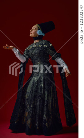 Elderly woman in renaissance attire gracefully raising hand while wearing modern headphones, immersed in music against deep red background. Elderly woman in renaissance attire gracefully raising hand while wearing modern headphones, immersed in music against deep red background. 123832547