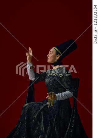 Elderly noblewoman in period dress elegantly raising fingers, as if making a grand gesture, golden chain in hand against deep red background. 123832554
