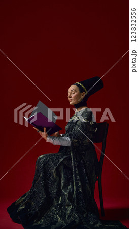 Noble elderly woman in renaissance-style dress intently reading book, hands delicately holding open pages against deep red background. 123832556