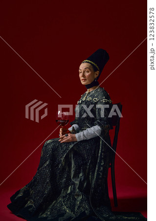 Elder noblewoman in renaissance attire holding large wine glass, elegantly sitting against rich red background with composed expression. 123832578