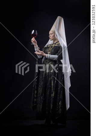Portrait of noblewoman dressed renaissance style elegantly holding large wine glass, analyzing deep red liquid against dark vintage background. 123832591
