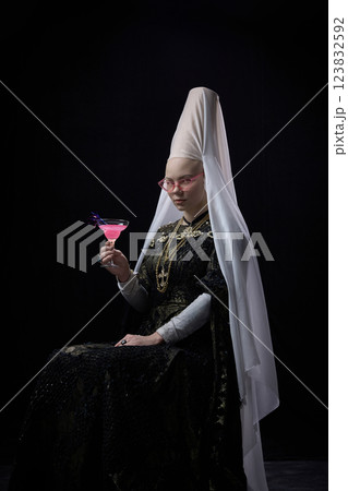 Young noblewoman in renaissance dress wearing pink sunglasses, holding martini glass with bright pink drink, gazing confidently against dark background. Young noblewoman in renaissance dress wearing pink sunglasses, holding martini glass with bright pink drink, gazing confidently against dark background. 123832592