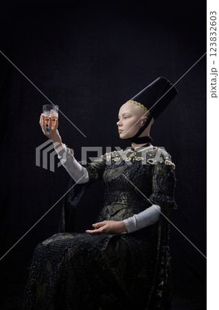 Portrait of young noblewoman dressed renaissance style clothes, holding glass of whiskey with focused expression against dark vintage background. Portrait of young noblewoman dressed renaissance style clothes, holding glass of whiskey with focused expression against dark vintage background. 123832603