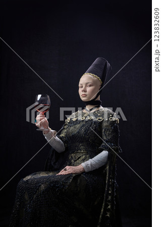 Young confident woman dressed renaissance style clothes, sitting with calm expression, holding glass of red wine against dark studio background. Young confident woman dressed renaissance style clothes, sitting with calm expression, holding glass of red wine against dark studio background. 123832609