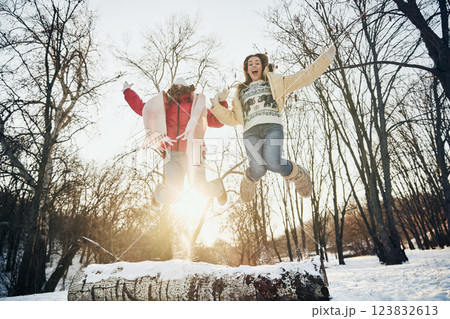 Two friends, women, dressed cozy and warm winter clothes, jumping excitedly in snowy park, arms raised in happiness. 123832613