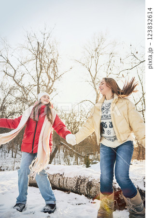 Two young women, dressed cozy winter outfits, holding hands, smiling as they play in snow-covered landscape. 123832614
