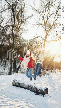 Two friends in warm jackets jumping over snowy log, enjoying outdoor adventure and having fun in snowy park. 123832615
