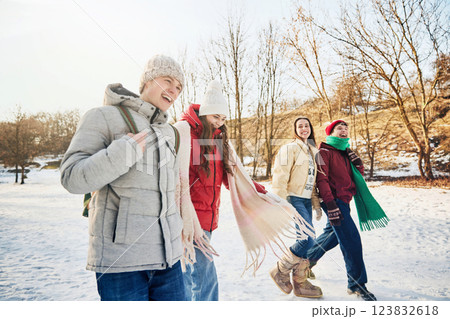 Four friends, men and women, in warm winter clothes walk together, laughing under golden sunlight in cold, winter park. Four friends, men and women, in warm winter clothes walk together, laughing under golden sunlight in cold, winter park. 123832618
