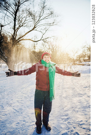 Joyful young man dressed in red hat and green scarf throws hands up, smiling while snowflakes fall in sunny park. Joyful young man dressed in red hat and green scarf throws hands up, smiling while snowflakes fall in sunny park. 123832628