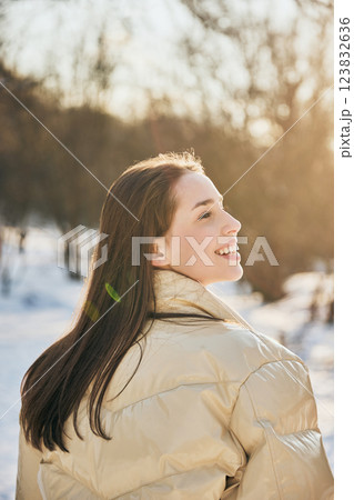 Joyful young woman dressed warm beige jacket smiles warmly in snowy outdoor setting. Rear view photo. Joyful young woman dressed warm beige jacket smiles warmly in snowy outdoor setting. Rear view photo. 123832636