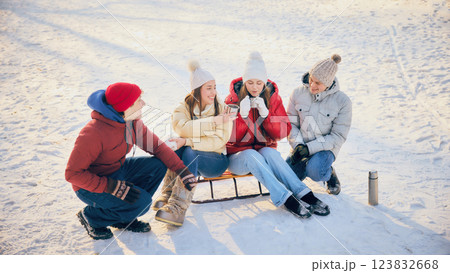 Group of friends, men and women, dressed comfy winter clothes, sitting in snow, sharing hot drinks, enjoying cozy winter moment together. Group of friends, men and women, dressed comfy winter clothes, sitting in snow, sharing hot drinks, enjoying cozy winter moment together. 123832668