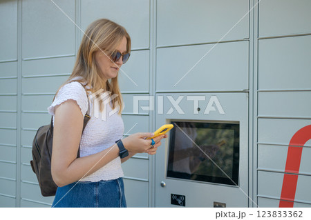 Cheerful young travel 30s woman using smart phone while standing near automatic lockers. New technologies in delivery service, self picking Cheerful young travel 30s woman using smart phone while standing near automatic lockers. New technologies in delivery service, self picking 123833362
