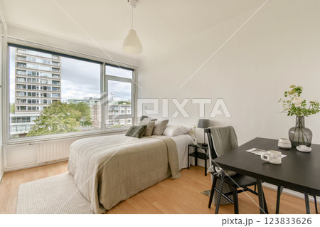 A bright modern bedroom showcasing a neatly made bed, stylish furniture, and large windows that offer a glimpse of the cityscape outside. 123833626