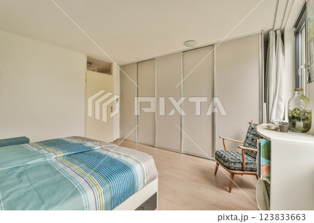 A stylish and contemporary bedroom featuring a neatly made bed with striped bedding, a cozy chair, and a sleek wardrobe, illuminated by natural light. A stylish and contemporary bedroom featuring a neatly made bed with striped bedding, a cozy chair, and a sleek wardrobe, illuminated by natural light. 123833663
