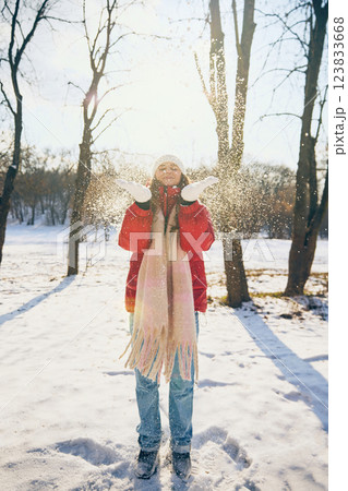 Young attractive woman wearing red comfy jacket and warm scarf, playfully tossing snow into air with happiness. Young attractive woman wearing red comfy jacket and warm scarf, playfully tossing snow into air with happiness. 123833668