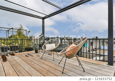 A modern rooftop patio featuring stylish woven chairs, wooden flooring, and a scenic view of the city skyline under a partly cloudy sky. 123833752
