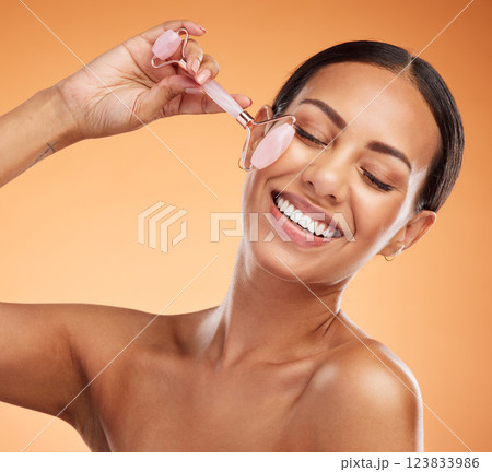 Jade roller, skincare and woman with massage for face beauty against an orange studio background. Happy, young and healthy model with a dermatology product for facial wellness and care with a smile Jade roller, skincare and woman with massage for face beauty against an orange studio background. Happy, young and healthy model with a dermatology product for facial wellness and care with a smile 123833986