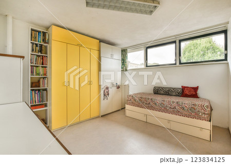 A contemporary bedroom showcasing a cozy bed with a vibrant patterned duvet, bright yellow cabinets, and ample natural light from large windows. A contemporary bedroom showcasing a cozy bed with a vibrant patterned duvet, bright yellow cabinets, and ample natural light from large windows. 123834125