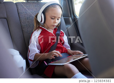 Child with tablet, headphones and safety belt in the car going home after soccer match, game or practice. Young girl in sports gear playing educational games online on tech device after training Child with tablet, headphones and safety belt in the car going home after soccer match, game or practice. Young girl in sports gear playing educational games online on tech device after training 123834381