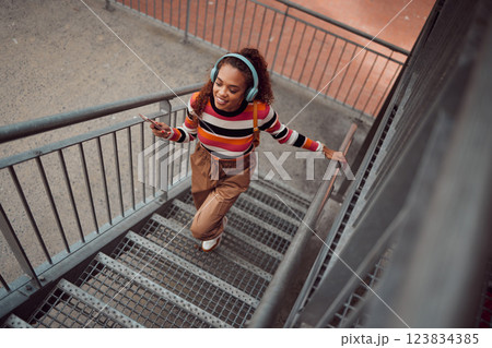 City, music and girl on stairs, phone in hand and listening to audio. Happy black woman with headphones streaming song, track or radio on smartphone in urban town, student freedom on weekend 123834385