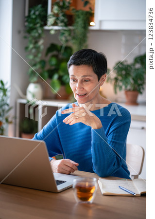 Focused mature woman gestures as explains tasks on video call at home, stting with tea and notes 123834736