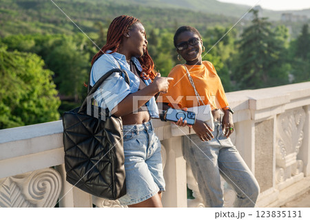 Laughing black friends chatting while lean on railing, waiting for taxi after booking on smartphone Laughing black friends chatting while lean on railing, waiting for taxi after booking on smartphone 123835131
