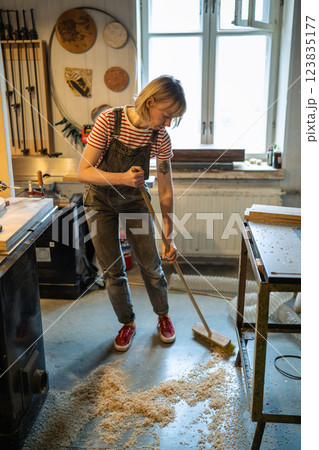 Carpenter woman cleaning up workshop after work. Handywoman sweep sawdust and ventilating for health 123835177